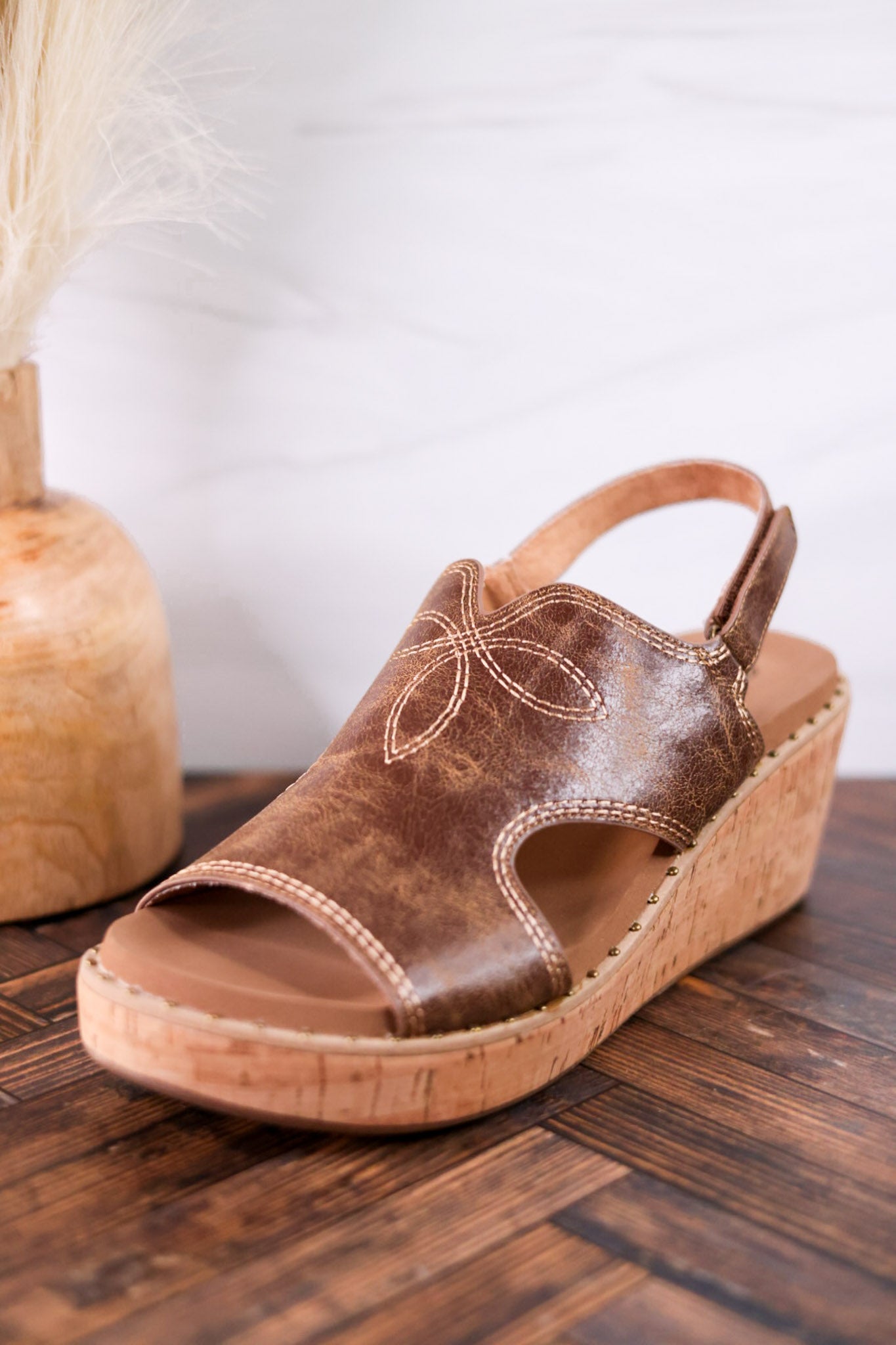 Cognac Faux Leather Wedges With Stitching featuring decorative floral stitch and cork wedge heel on wooden floor background