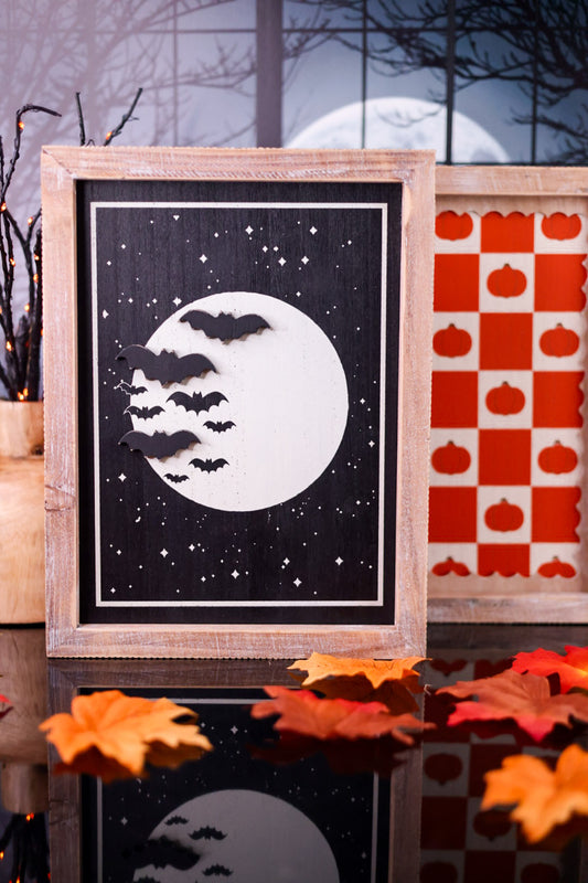 Framed Halloween-themed artwork with bats and moon against a night sky, displayed on a surface with autumn leaves.