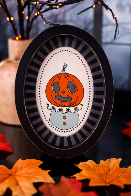 Halloween-themed pumpkin decoration in a black frame with autumn leaves and lights in the background.