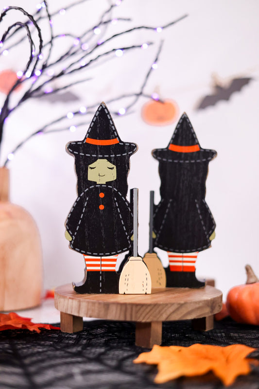 Two witch figurines on a wooden stand with Halloween decorations in the background.