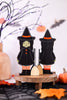 Two witch figurines on a wooden stand with Halloween decorations in the background.