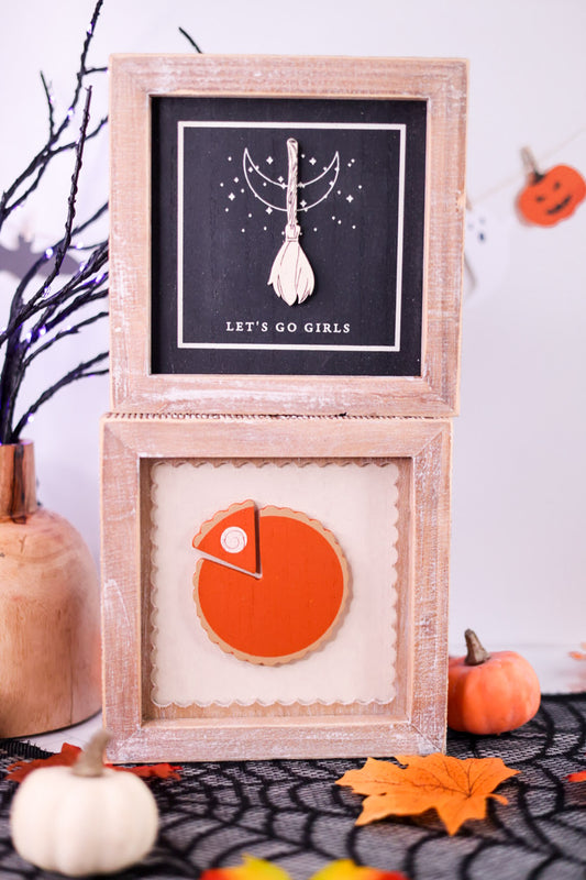 Two framed artworks on a Halloween-themed table with pumpkins and leaves.