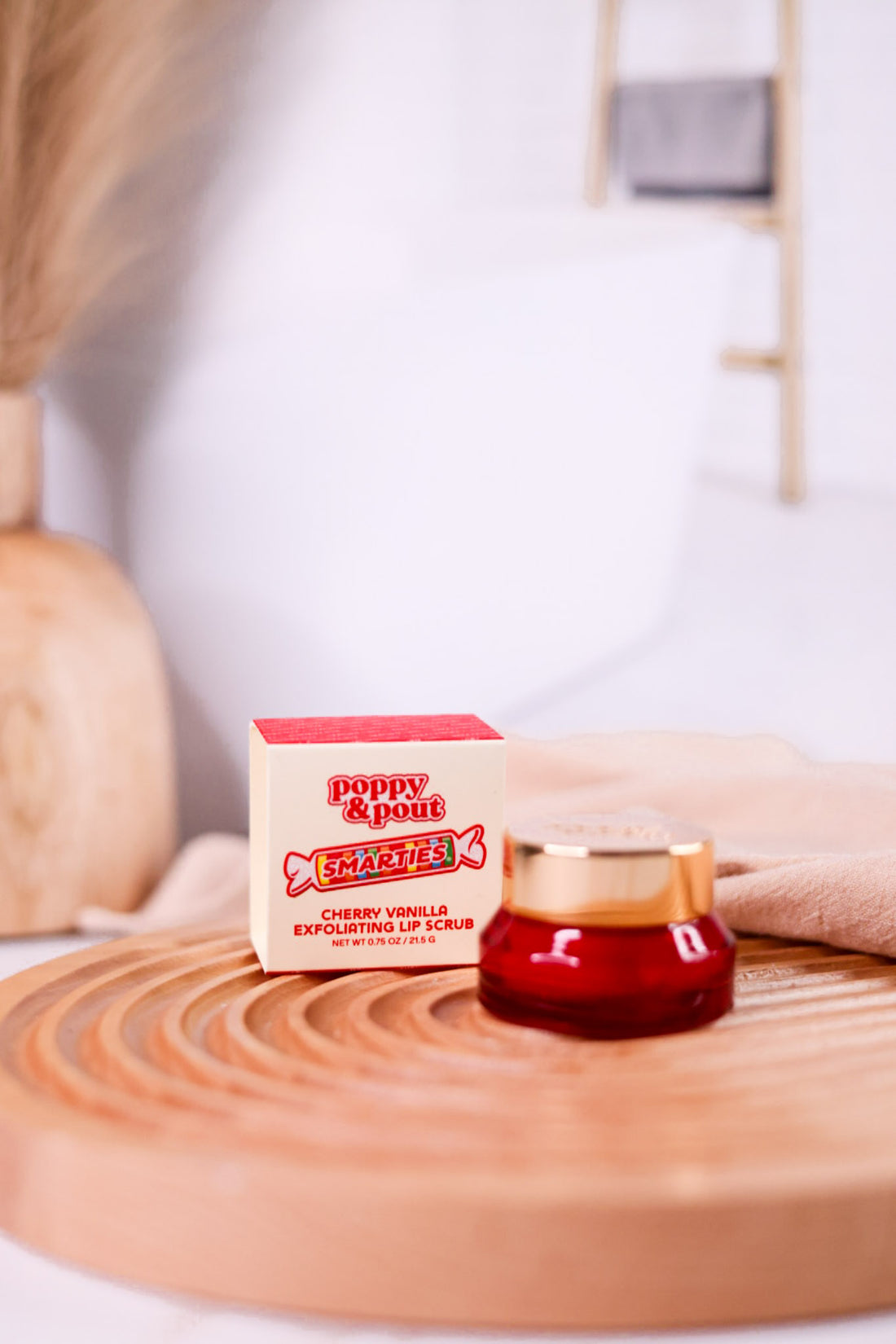 Smarties Exfoliating Lip Scrub in red jar with gold lid displayed next to branded box on wooden surface
