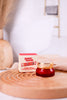 Smarties Exfoliating Lip Scrub in red jar with gold lid displayed next to branded box on wooden surface