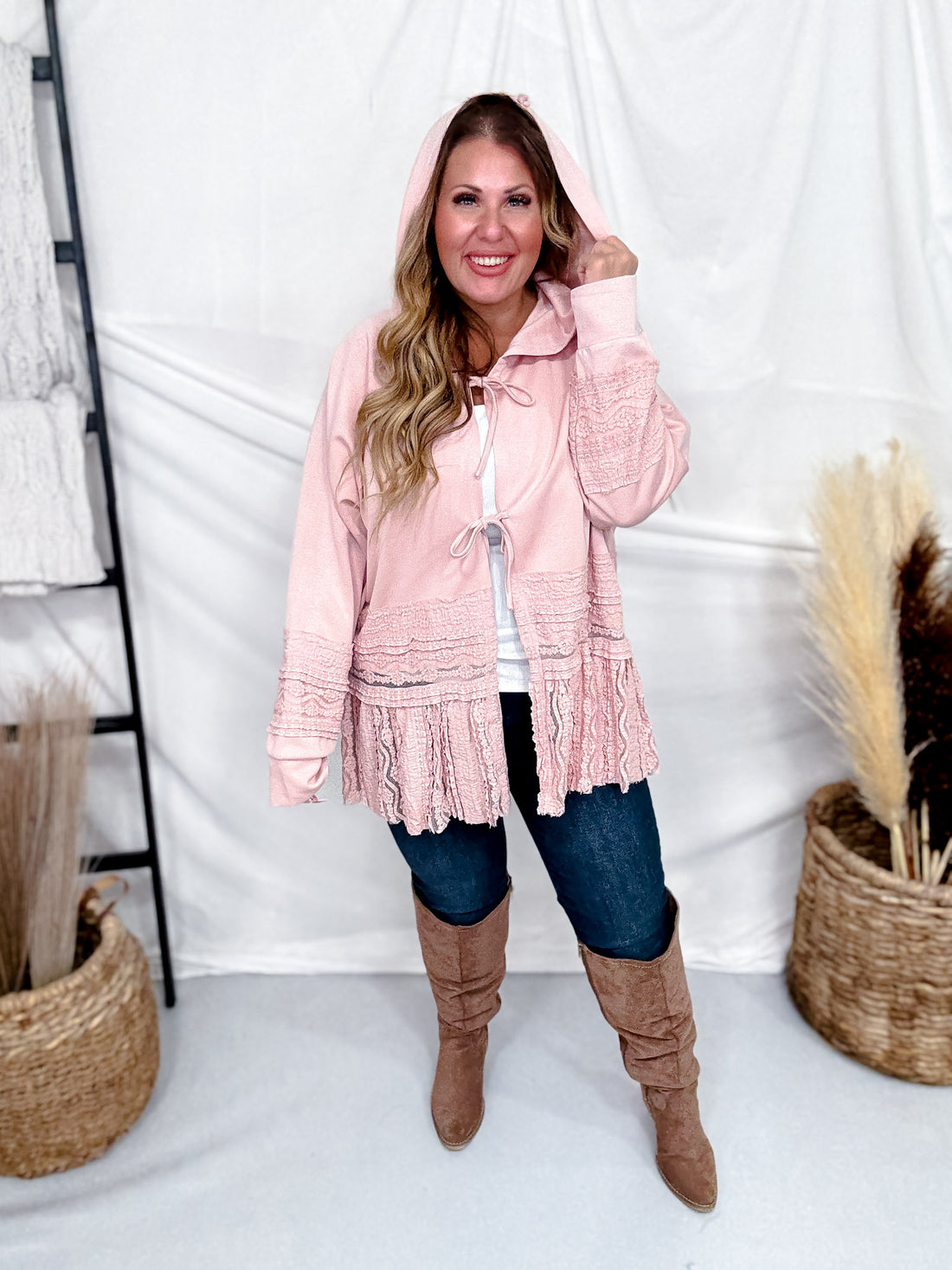 Woman wearing Dusty Pink Laced Front Tie Hooded Jacket Cardigan with jeans and tan suede boots indoors smiling