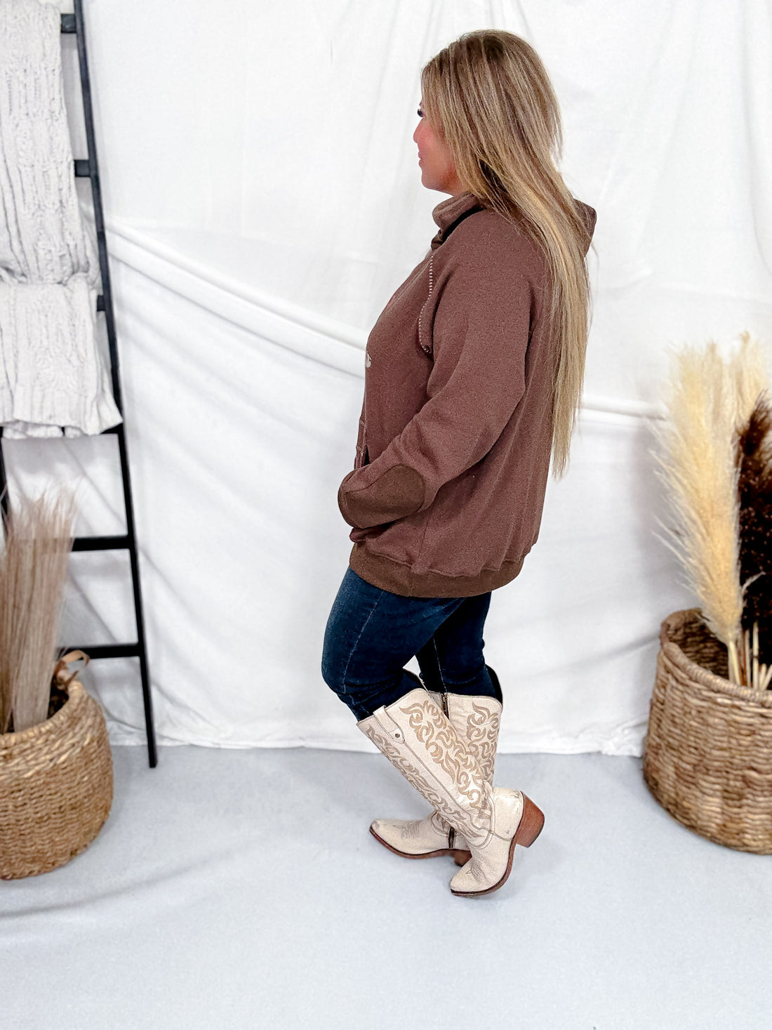 Side view of woman wearing Western Bullrider Hoodie In Chocolate Brown with dark jeans and embroidered cream boots