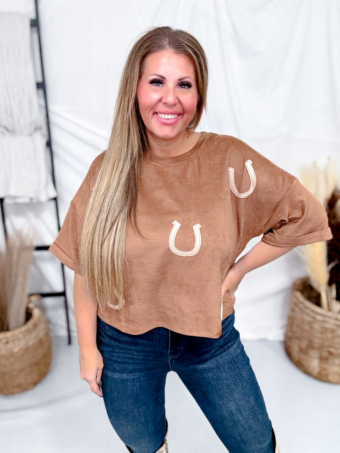 Smiling woman wearing Horseshoe Patch Faux Suede Top in warm brown paired with dark jeans in bright indoor setting