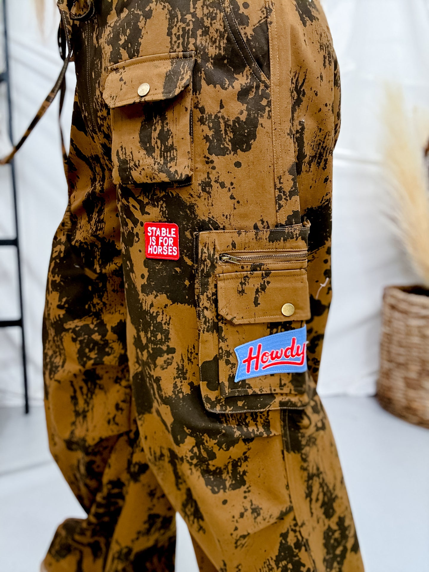 Close-up of Brown Cow Camo Print Cargo Pants With Western Patches featuring pockets and embroidered patches in neutral setting