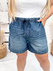 Close-up of woman wearing Judy Blue High Waist Featherweight Shorts With Drawstring Tie in mid wash denim with hands in pockets