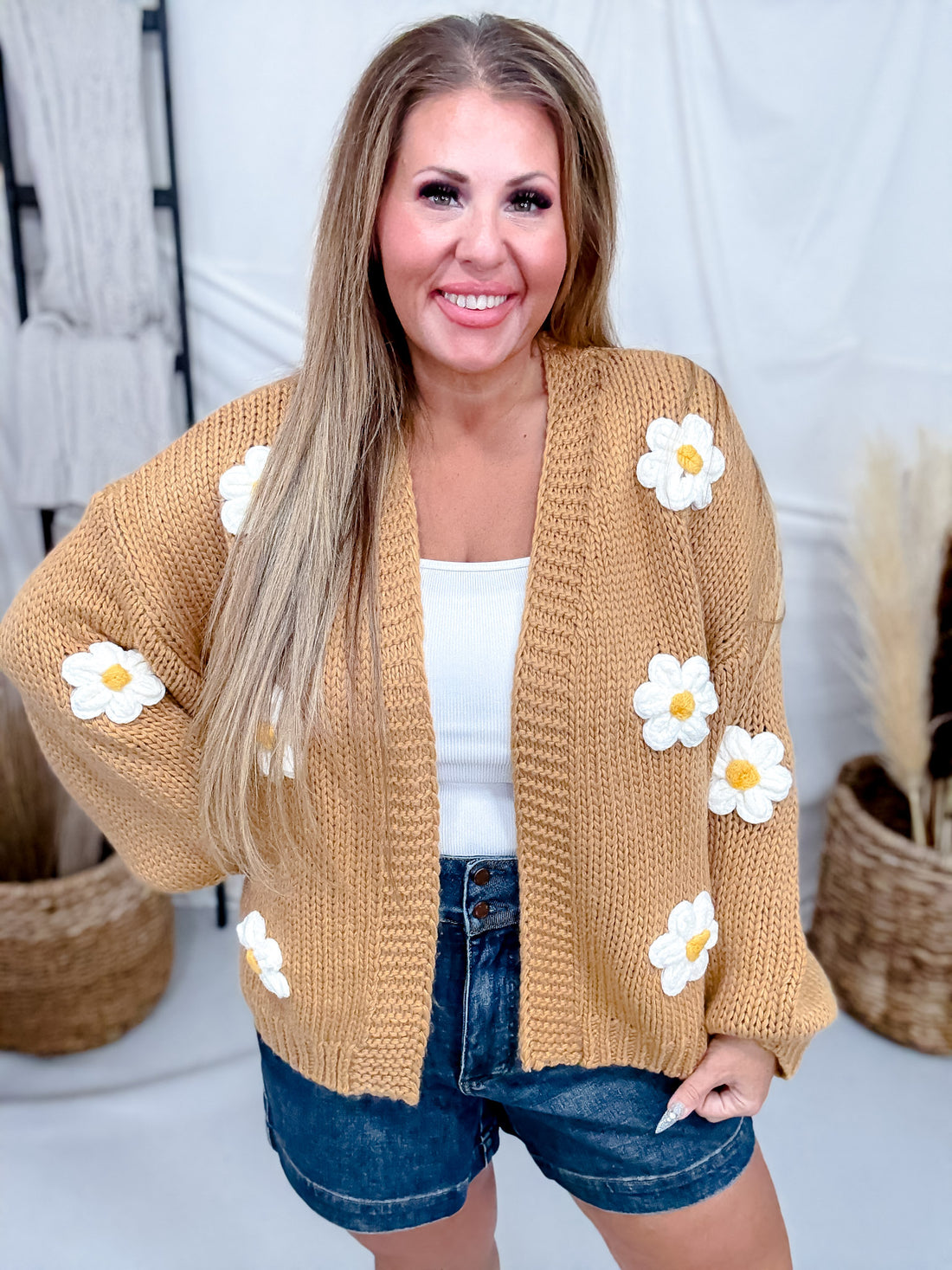 Smiling woman wearing Knit Cardigan with Daisies in Camel Brown paired with white tank top and denim shorts in cozy setting