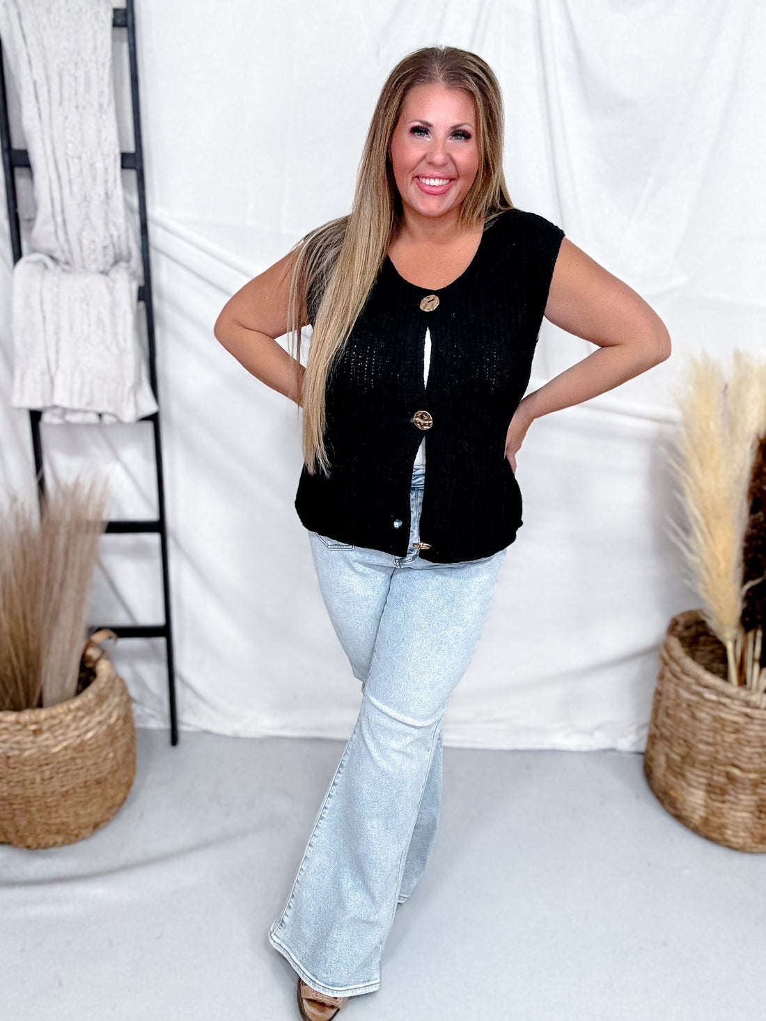 Woman smiling wearing Black Sleeveless Crochet Sweater Vest With Metallic Buttons paired with light wash flared jeans indoors