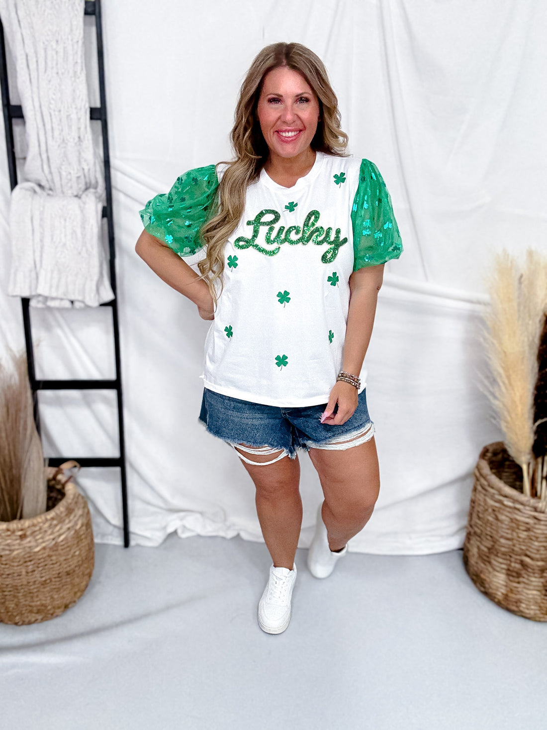 Smiling woman wearing Sequined "Lucky" Top With Tulle Sleeves with green shamrocks paired with distressed denim shorts indoors